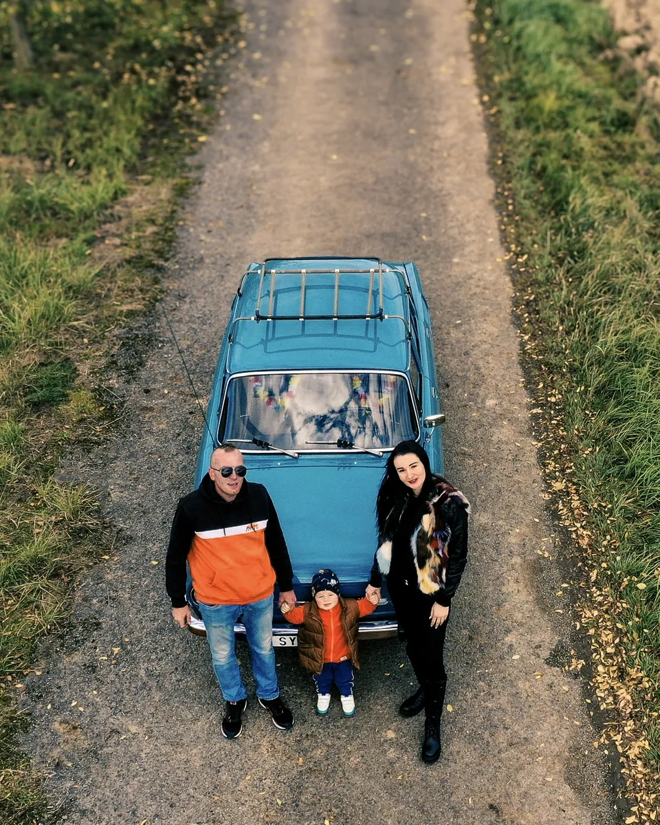 Rodina stojí na cestě před modrým autem, fotografováno shora